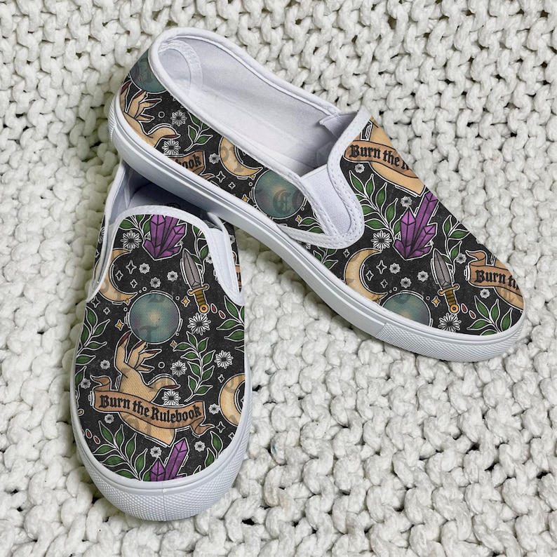 K&ouml;nnte beinhalten: Paar Slip-On-Schuhe mit schwarzem Hintergrund und einem sich wiederholenden Muster aus Monden, Kristallen, Dolchen und H&auml;nden. Die Schuhe haben wei&szlig;e Sohlen und den Text "Burn the Rulebook" auf einem Banner.