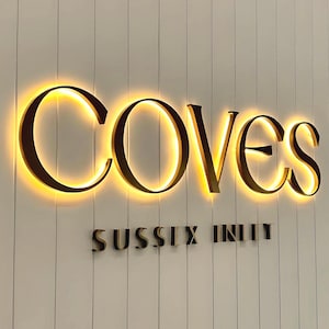 May include: A wall-mounted sign featuring the word COVES in large, dark, backlit letters that emit a warm golden glow. Below this, the words SUSSEX INLET appear in smaller, dark, non-illuminated letters against a white vertical paneled wall.