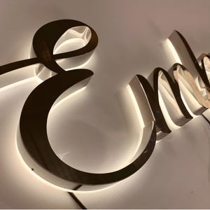 May include: A three-dimensional sign featuring the cursive letter E followed by the letters n and m in a polished metallic finish. The letters are mounted with a backlit LED glow that casts a soft white light onto the surface behind the sign.