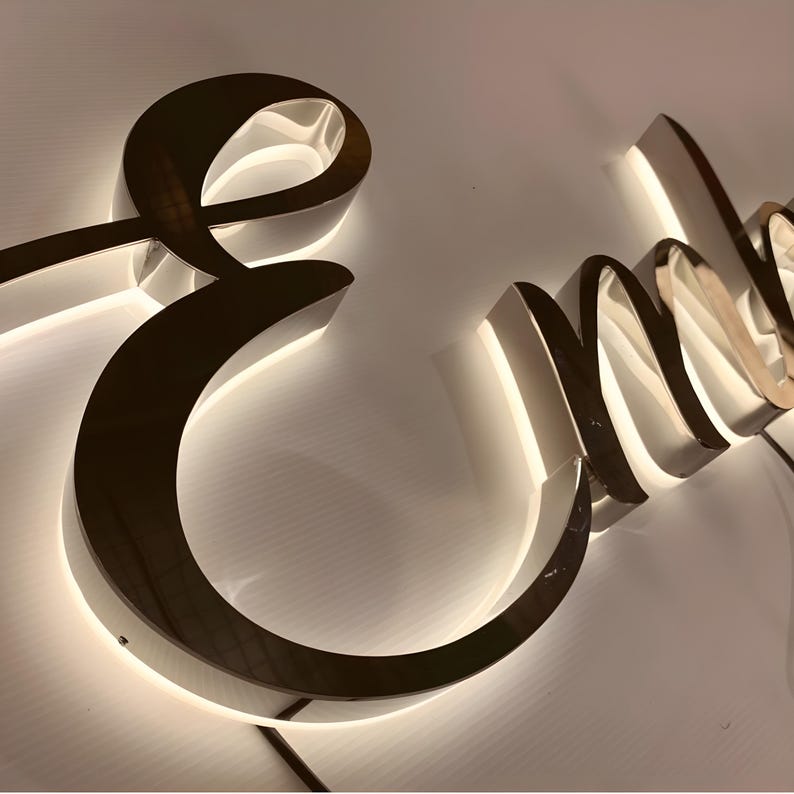 May include: A three-dimensional sign featuring the cursive letter E followed by the letters n and m in a polished metallic finish. The letters are mounted with a backlit LED glow that casts a soft white light onto the surface behind the sign.