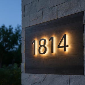 K&ouml;nnte beinhalten: Beleuchtete Hausnummern, "1814", sind auf einer dunklen Holztafel montiert. Die schwarzen Ziffern werden von einem warmen Licht hinterleuchtet. Die Tafel ist an einer Steinmauer befestigt.