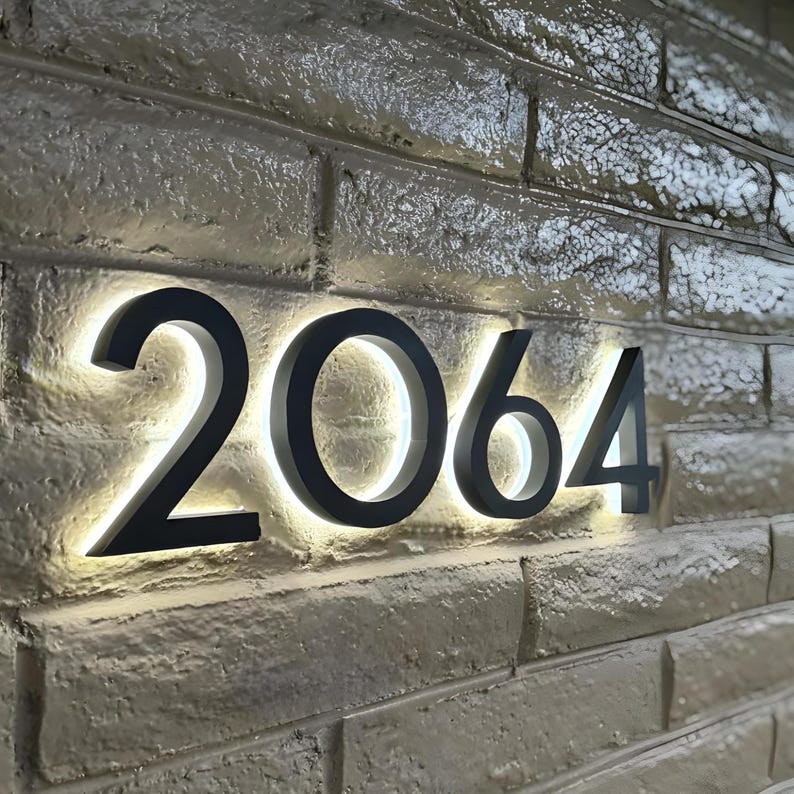 Peut inclure: Num&eacute;ros de maison noirs &eacute;clair&eacute;s 2064 mont&eacute;s sur un mur de briques textur&eacute;. Les chiffres sont r&eacute;tro-&eacute;clair&eacute;s avec une lumi&egrave;re blanche chaude, cr&eacute;ant un look moderne. Le mur de briques est gris clair.