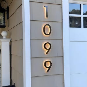 Peut inclure: Num&eacute;ros de maison modernes, 1099, avec un design r&eacute;tro-&eacute;clair&eacute;. Les chiffres noirs sont mont&eacute;s sur un mur clair. La lueur chaude derri&egrave;re les chiffres cr&eacute;e un effet visuel &eacute;l&eacute;gant et lumineux.