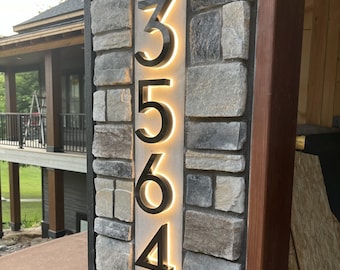 May include: A modern address sign with the numbers 3564 illuminated against a light-colored background. The numbers are black and mounted on a stone wall. The sign is rectangular and vertically oriented.