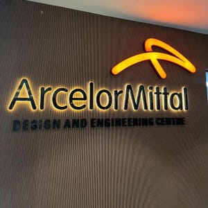 May include: A wall-mounted sign featuring the ArcelorMittal logo in glowing orange, with the company name ArcelorMittal in backlit black lettering, and the text DESIGN AND ENGINEERING CENTRE in black below, all set against a vertical wood-slat wall.