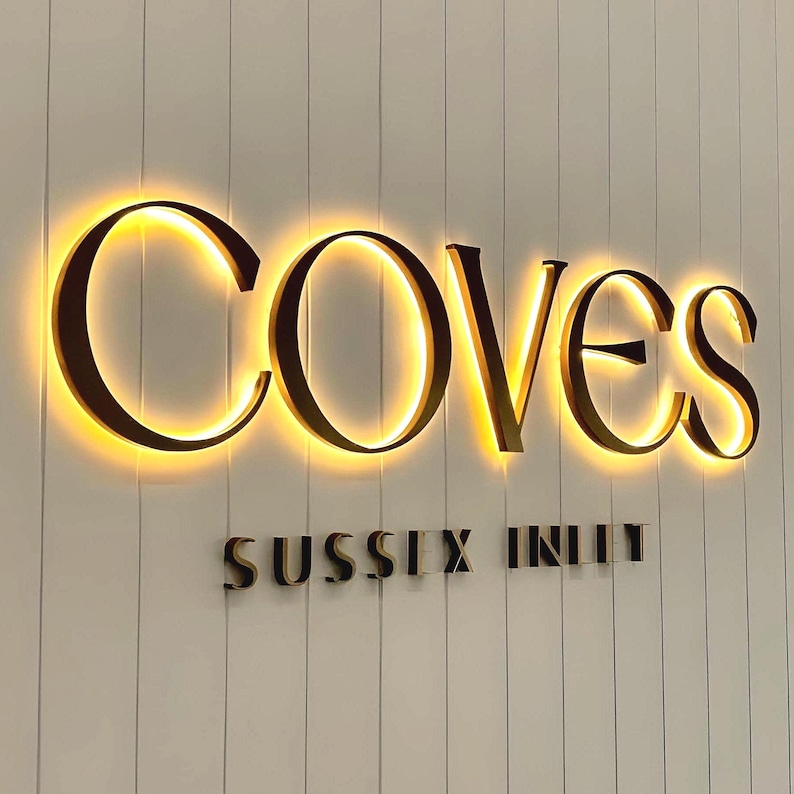 May include: A wall-mounted sign featuring the word COVES in large, dark, backlit letters that emit a warm golden glow. Below this, the words SUSSEX INLET appear in smaller, dark, non-illuminated letters against a white vertical paneled wall.