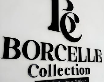 May include: Black acrylic sign with the Borcelle Collection logo and text on a white wall. The logo is a stylized "Bc" and the text reads "Borcelle Collection" and "Live Bodly, Dress Modesty."