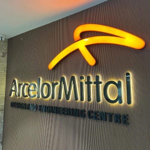 May include: A wall-mounted sign featuring the ArcelorMittal logo in glowing orange, with the company name and the text DESIGN AND ENGINEERING CENTRE in black, backlit lettering against a vertical, dark brown, ribbed textured wall panel.