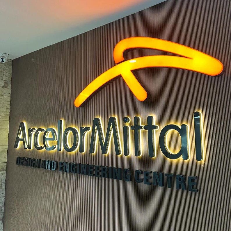 May include: A wall-mounted sign featuring the ArcelorMittal logo in glowing orange, with the company name and the text DESIGN AND ENGINEERING CENTRE in black, backlit lettering against a vertical, dark brown, ribbed textured wall panel.