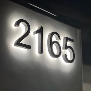 Peut inclure: Num&eacute;ros d'adresse en m&eacute;tal noir &eacute;clair&eacute;s, 2165, mont&eacute;s sur un mur gris clair. Les chiffres sont r&eacute;tro-&eacute;clair&eacute;s par une lumi&egrave;re blanche vive, cr&eacute;ant un contraste saisissant. Police sans-serif.