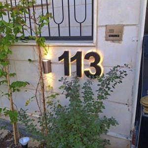 Peut inclure: Num&eacute;ro de maison &eacute;clair&eacute; 113 en noir, dans une police moderne, mont&eacute; sur un mur textur&eacute;. Les chiffres ont un r&eacute;tro&eacute;clairage chaleureux. Un petit luminaire ext&eacute;rieur est &agrave; gauche des chiffres, avec de la verdure au premier plan.