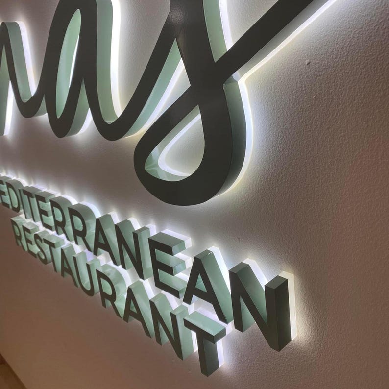 May include: A three-dimensional wall sign featuring dark-colored lettering with a bright white LED backlight. The sign displays the text "MEDITERRANEAN RESTAURANT" in a bold, sans-serif font, positioned below a larger, cursive script word.