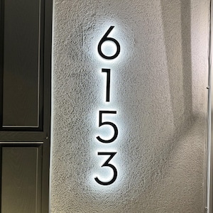 Peut inclure: Numéros de maison noirs éclairés 6153 montés sur un mur gris clair texturé. Les chiffres sont dans une police moderne sans empattement, avec une lueur blanche brillante émanant de derrière chaque chiffre. Les chiffres sont alignés verticalement.