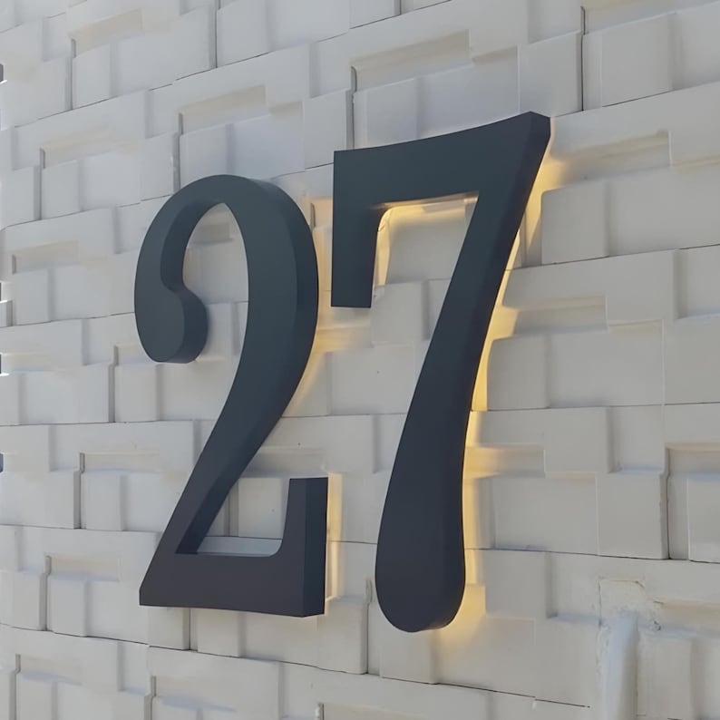 Peut inclure: Le chiffre 27 gris fonc&eacute; est mont&eacute; sur un mur blanc textur&eacute;. Les chiffres sont grands et stylis&eacute;s, avec une lumi&egrave;re chaude &eacute;manant de derri&egrave;re eux, cr&eacute;ant une lueur subtile. Le mur pr&eacute;sente un motif r&eacute;p&eacute;titif de petits blocs carr&eacute;s.