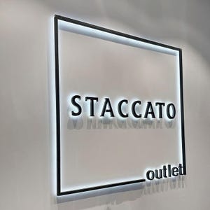 May include: A square wall-mounted sign featuring a black metal frame with integrated white LED backlighting. The sign displays the word STACCATO in large, bold, black capital letters, with the word outlet in smaller black letters in the bottom corner.