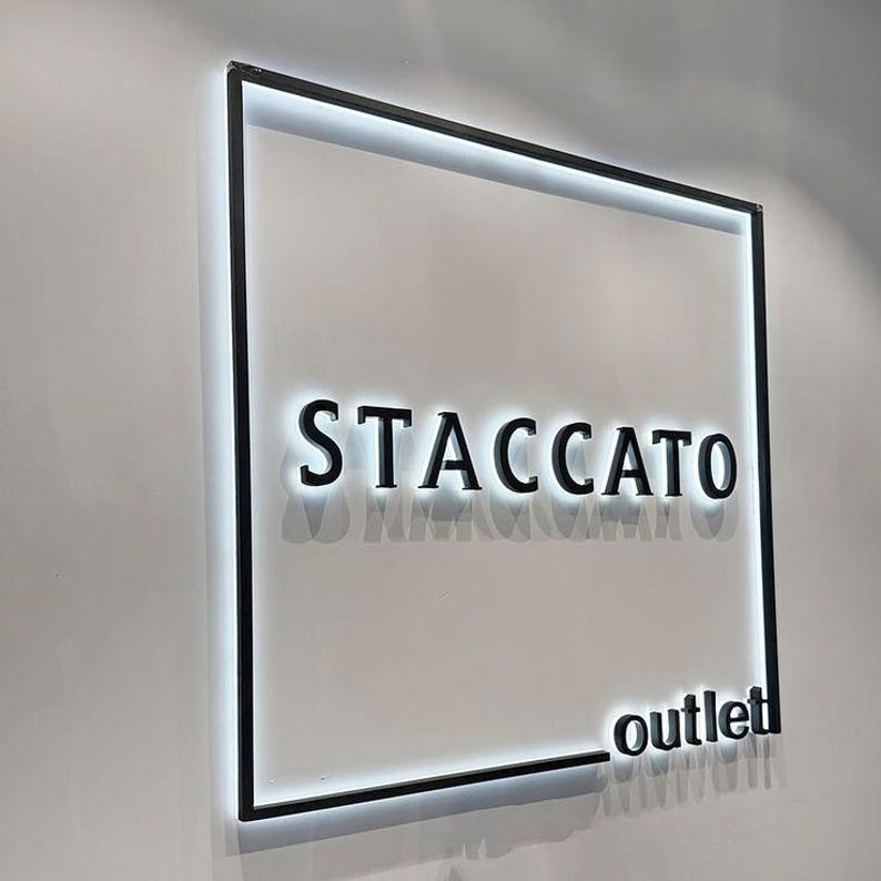 May include: A square wall-mounted sign featuring a black metal frame with integrated white LED backlighting. The sign displays the word STACCATO in large, bold, black capital letters, with the word outlet in smaller black letters in the bottom corner.