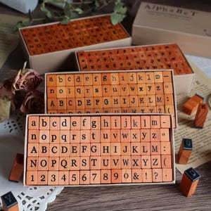 May include: A set of wooden rubber stamps arranged in rectangular boxes, featuring lowercase and uppercase letters, numbers, and punctuation marks. The stamps are light brown with black ink characters. The box lid is labeled with the text AlPhAbEt.