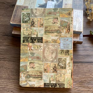 May include: A vintage-style journal with a collage of antique labels and advertisements. The cover features a variety of images and text, including "Sunlight Soap" and "British Empire". The journal is resting on a wooden surface, with other books visible.