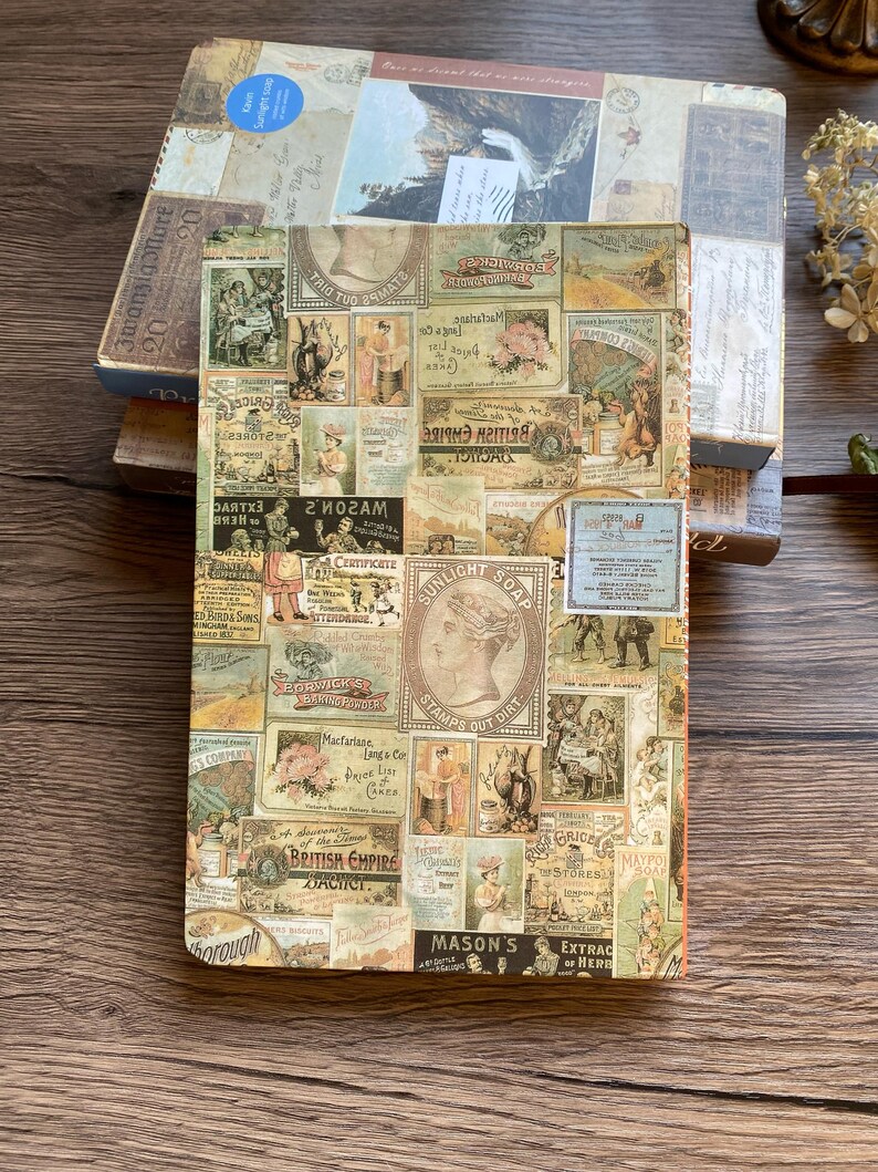 May include: A vintage-style journal with a collage of antique labels and advertisements. The cover features a variety of images and text, including "Sunlight Soap" and "British Empire". The journal is resting on a wooden surface, with other books visible.