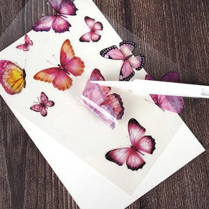 Puede incluir: Una hoja de pegatinas de mariposas en varios tama&ntilde;os y tonos de rosa, morado y amarillo. Las pegatinas est&aacute;n sobre un respaldo transparente, con un papel blanco debajo. Una herramienta blanca est&aacute; despegando una pegatina de la hoja.