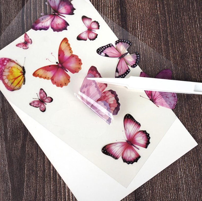 Puede incluir: Una hoja de pegatinas de mariposas en varios tama&ntilde;os y tonos de rosa, morado y amarillo. Las pegatinas est&aacute;n sobre un respaldo transparente, con un papel blanco debajo. Una herramienta blanca est&aacute; despegando una pegatina de la hoja.