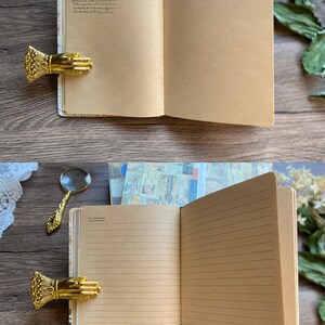 May include: Open journal with lined pages and decorative edges. The journal is held open by a gold-colored hand-shaped clip. A magnifying glass rests nearby. The journal's pages are a light brown color.