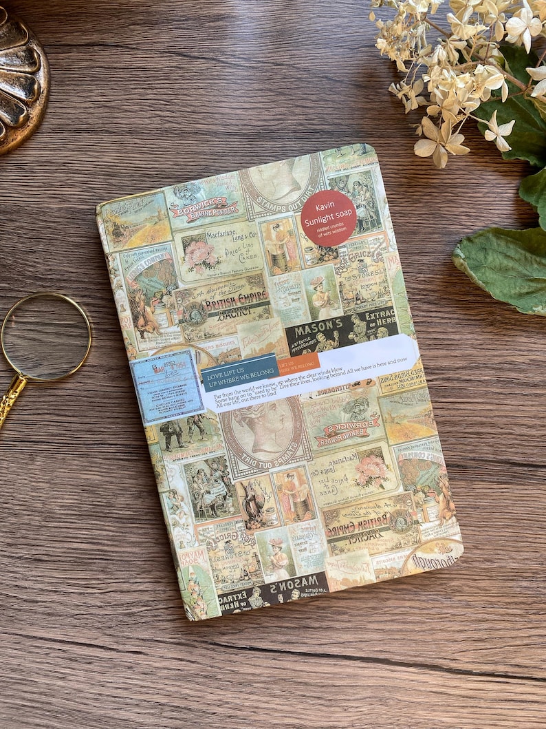 May include: A rectangular notebook with a collage of vintage-style labels and stamps. The notebook has a sticker that reads "Kavin Sunlight soap." The background is a wooden surface. The notebook is a unique stationery item.