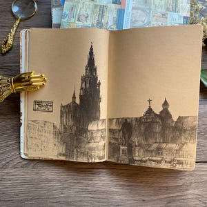 May include: An open journal with brown pages features a detailed pen and ink drawing of a cathedral and city buildings. A gold-colored hand-shaped page holder and a magnifying glass are visible.