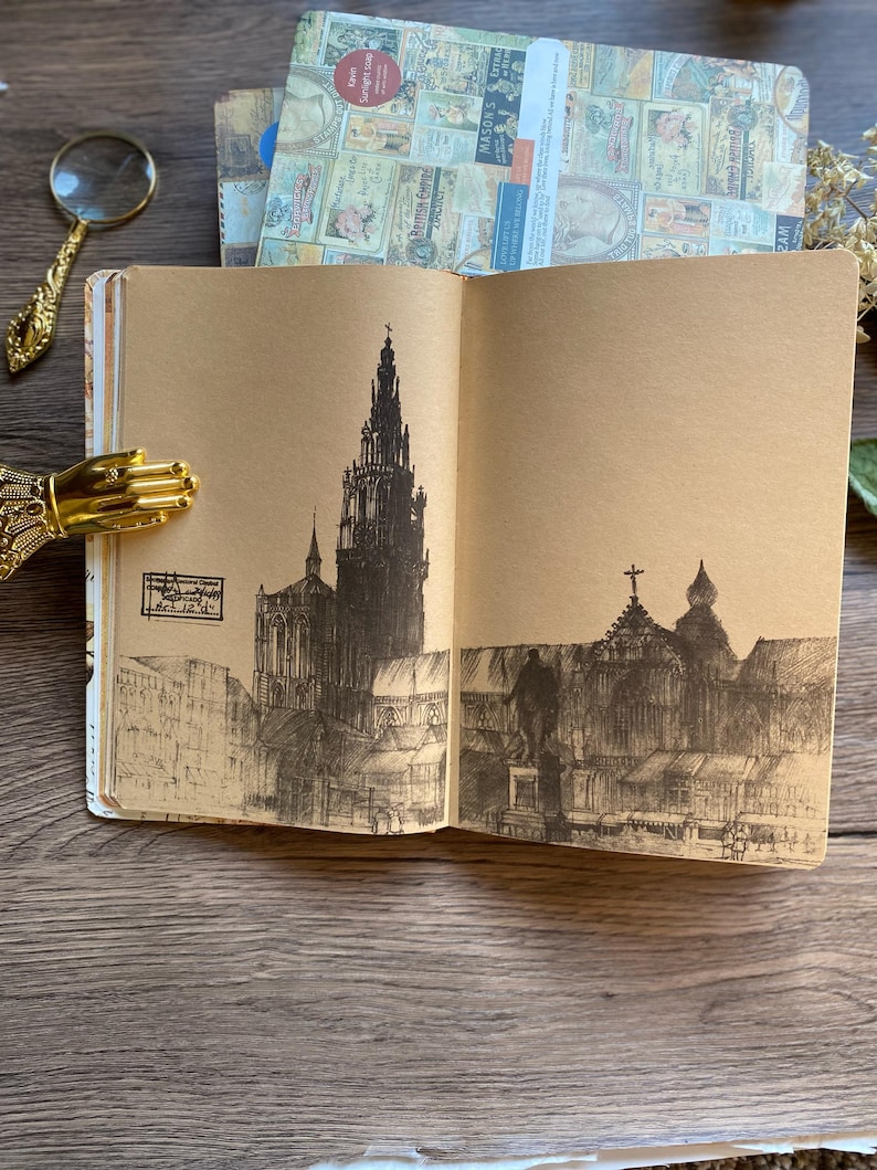 May include: An open journal with brown pages features a detailed pen and ink drawing of a cathedral and city buildings. A gold-colored hand-shaped page holder and a magnifying glass are visible.
