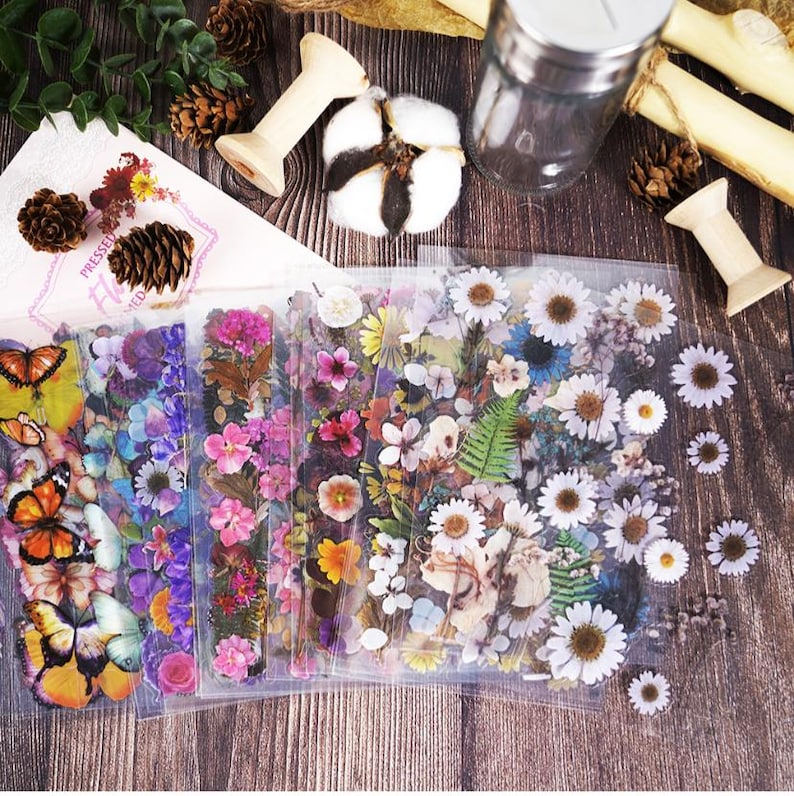 Puede incluir: Pegatinas florales con mariposas y diversas flores en tonos rosa, morado, amarillo y blanco. Las pegatinas est&aacute;n en un embalaje de pl&aacute;stico transparente, con elementos decorativos como pi&ntilde;as y algod&oacute;n.