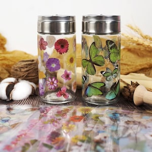 Puede incluir: Dos frascos de vidrio transparente con tapas plateadas. Uno est&aacute; decorado con calcoman&iacute;as de flores coloridas, el otro con calcoman&iacute;as de mariposas verdes. Los frascos est&aacute;n sobre una superficie de madera con elementos decorativos en el fondo.