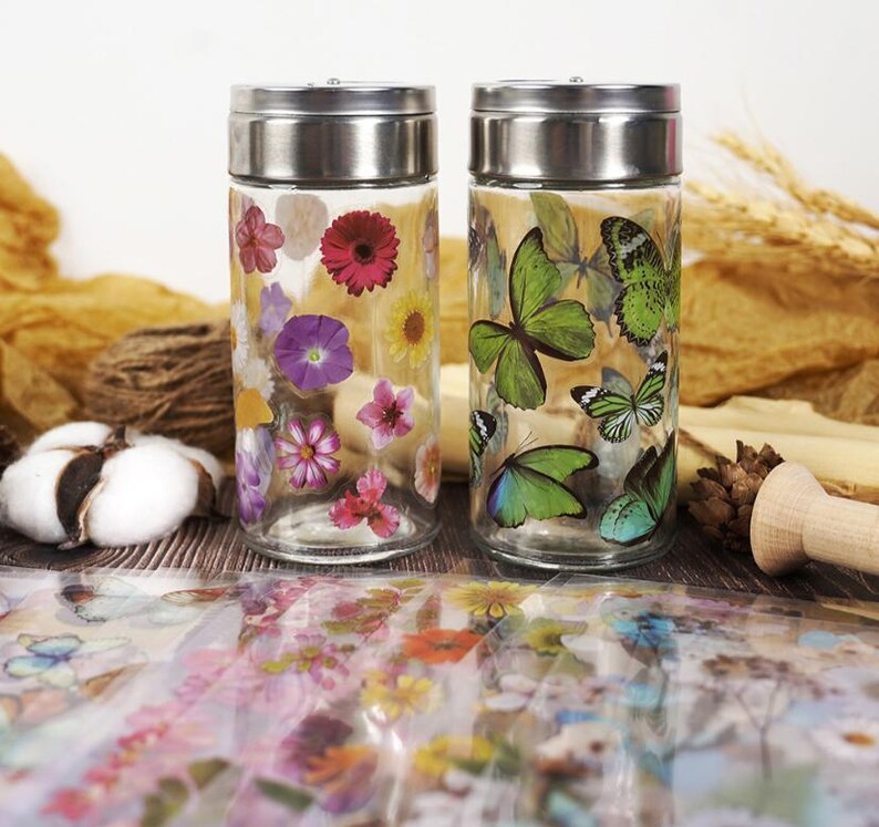 Puede incluir: Dos frascos de vidrio transparente con tapas plateadas. Uno est&aacute; decorado con calcoman&iacute;as de flores coloridas, el otro con calcoman&iacute;as de mariposas verdes. Los frascos est&aacute;n sobre una superficie de madera con elementos decorativos en el fondo.
