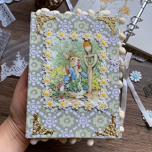 May include: A decorative journal with a floral and bunny illustration. The cover is light green with a daisy pattern and gold corner accents. White pom-poms line the edges, and a daisy-trimmed frame surrounds the central image.