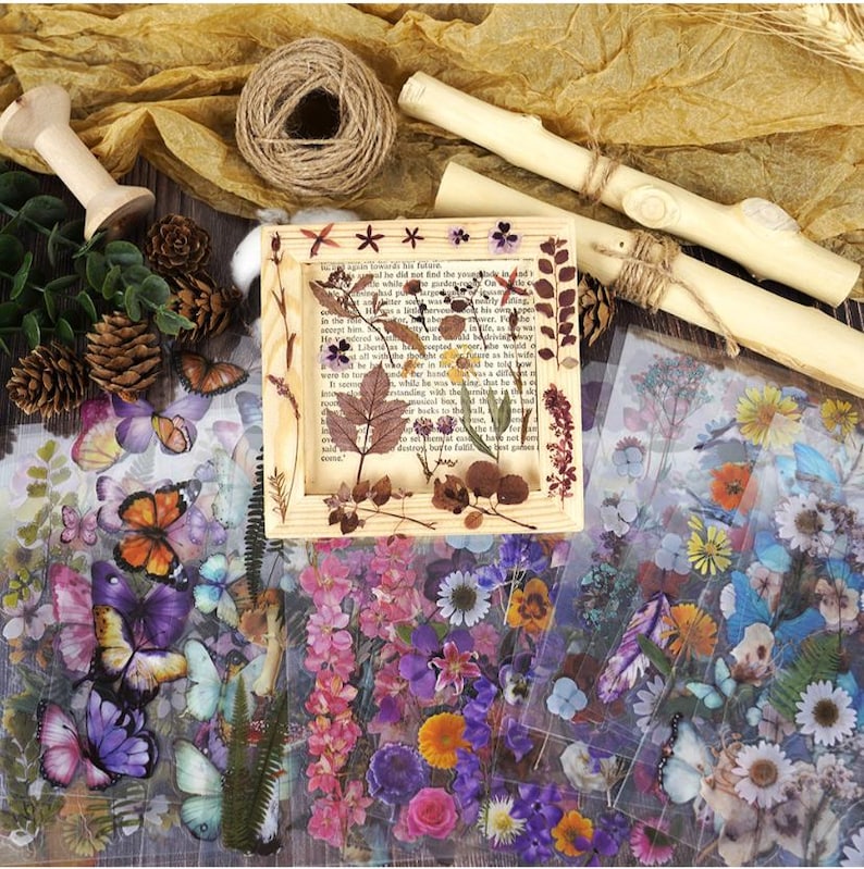 Puede incluir: Una colecci&oacute;n de materiales de manualidades que incluye un marco de madera con flores y hojas secas, pegatinas de mariposas y flores, hilo, clavijas de madera y pi&ntilde;as. La paleta de colores incluye tonos naturales de marr&oacute;n, verde y amarillo, con toques de morado y naranja.