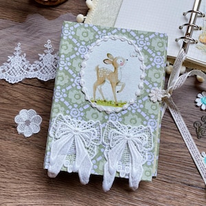 May include: A small, handmade journal with a green floral pattern cover. The cover features a circular embroidered image of a fawn. White lace bows and a ribbon closure add decorative details. The journal is a charming, handcrafted item.