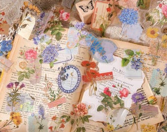 Puede incluir: Una colección de pegatinas florales y efímeras de estilo vintage, con rosas, hortensias y mariposas, dispuestas sobre un libro abierto. Las pegatinas translúcidas presentan una variedad de colores y diseños, perfectas para scrapbooking y proyectos de manualidades.
