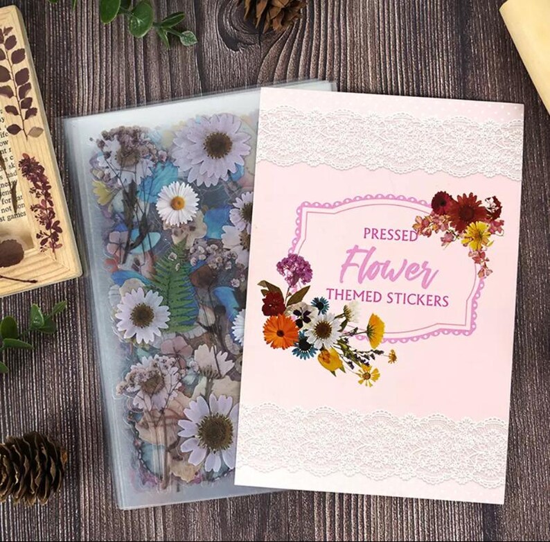 Puede incluir: Una funda de pl&aacute;stico transparente llena de flores prensadas y una tarjeta rosa con el texto "PRESSED Flower THEMED STICKERS". La tarjeta tiene un borde de encaje blanco y un dise&ntilde;o floral.