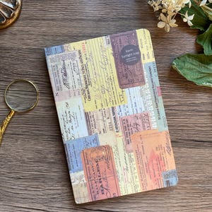May include: A rectangular journal with a collage of vintage ticket stubs and ephemera. The cover features a variety of colors, including yellow, orange, blue, and beige. The text "Kavin Sunlight soap" is visible on a circular sticker. The journal is on a wooden surface.