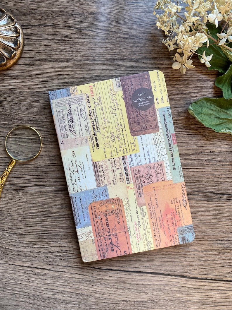 May include: A rectangular journal with a collage of vintage ticket stubs and ephemera. The cover features a variety of colors, including yellow, orange, blue, and beige. The text "Kavin Sunlight soap" is visible on a circular sticker. The journal is on a wooden surface.