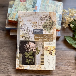 May include: A collection of vintage-style journals with collage designs. The top journal displays a floral and stamp motif with text that reads "Strange breeze is too gentle like as old friced like old tinca." The journals are on a wooden surface.