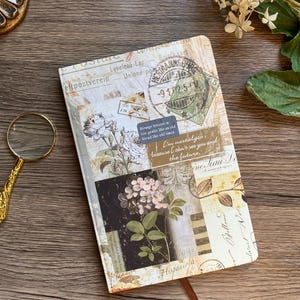 May include: A decorative journal with a vintage postcard design. The cover features floral illustrations, postage stamps, and handwritten script. A brown ribbon bookmark is visible. The text on the cover includes "Strange breeze is too gentle like as old friend like old times."
