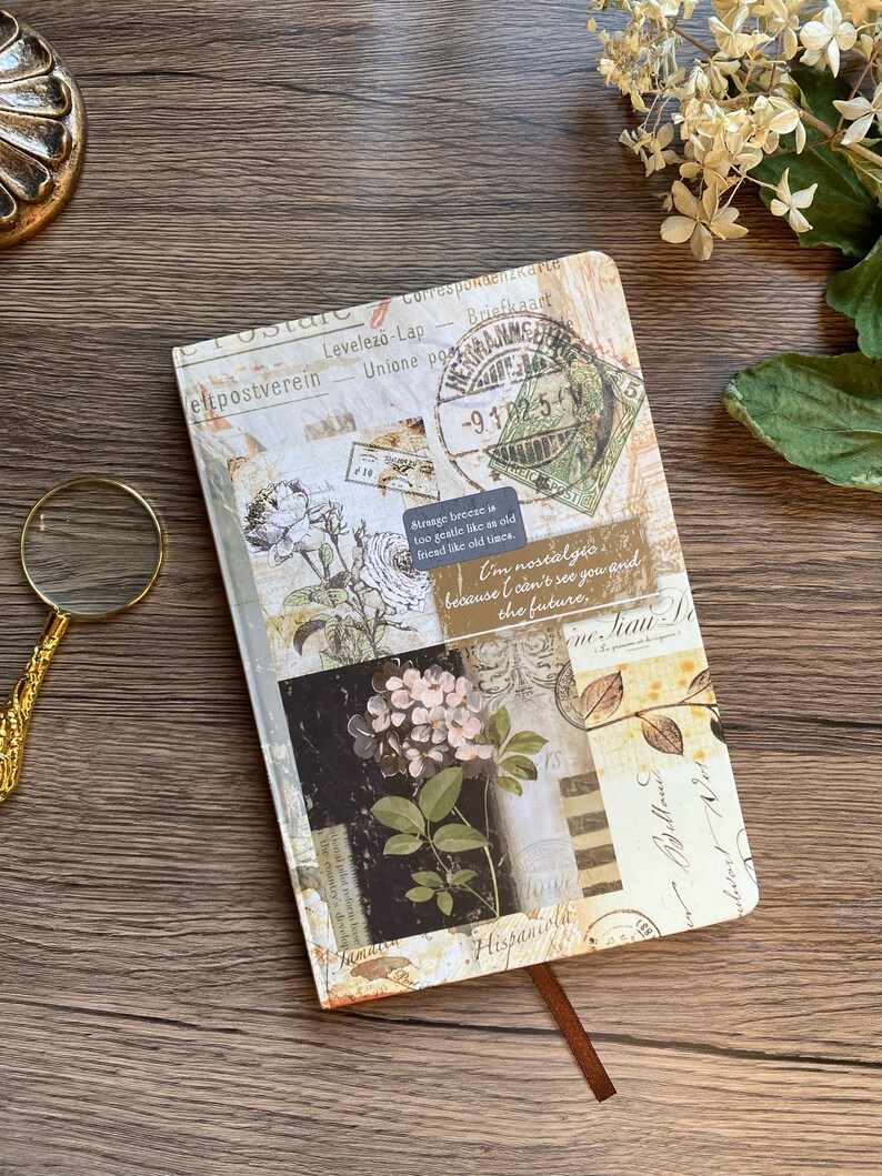 May include: A decorative journal with a vintage postcard design. The cover features floral illustrations, postage stamps, and handwritten script. A brown ribbon bookmark is visible. The text on the cover includes "Strange breeze is too gentle like as old friend like old times."