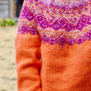 May include: A long-sleeved knit sweater featuring a vibrant orange base color. The yoke displays a detailed circular pattern in shades of bright pink, light pink, and soft orange, finished with a ribbed crew neckline and textured knit stitches.