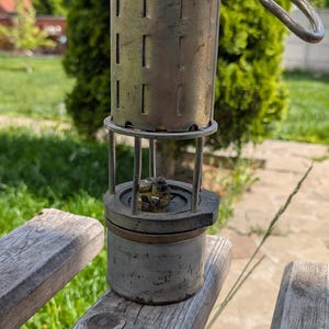May include: An antique brass miner's lamp with a cylindrical body and a wire handle. The lamp has a perforated top and a glass enclosure. It is sitting on a wooden bench outdoors.