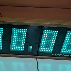 Pode incluir: Um relógio digital vintage com moldura de madeira exibe a hora 11:11 em luzes LED verdes brilhantes. O mostrador do relógio é preto e o texto "Электроника 7" é visível na parte inferior. O relógio é retangular.