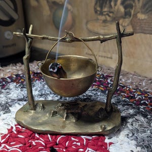 May include: A brass-colored metal incense burner shaped like a miniature cauldron hanging from a tripod stand. The stand features two branch-like vertical supports on an oval base. A single stick of incense burns inside the bowl, emitting smoke.