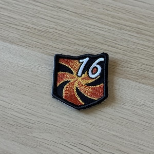 Puede incluir: Un parche negro con forma de escudo, con el número "76" en blanco y un diseño estilizado en naranja y amarillo. El parche tiene un borde negro y es probablemente un emblema o insignia.