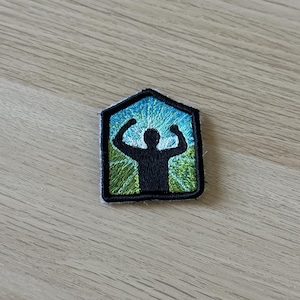 Puede incluir: Parche bordado con una silueta negra de una persona con los brazos levantados en señal de celebración. La figura se encuentra sobre un cielo azul con un efecto de luz radiante y hierba verde. El parche está enmarcado por un borde negro.