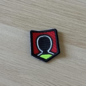 Op de afbeelding: Geborduurde patch met een schildontwerp, rode achtergrond en zwarte omtrek. Het heeft een witte silhouet van een hoofd en schouders, met een neon groen gedeelte aan de onderkant. De patch is ongeveer 5 cm breed.