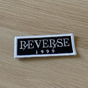 Puede incluir: Un parche rectangular negro con texto bordado en blanco que dice "REVERSE" encima de "1999". El parche tiene un borde blanco y está sobre una superficie de madera marrón claro.